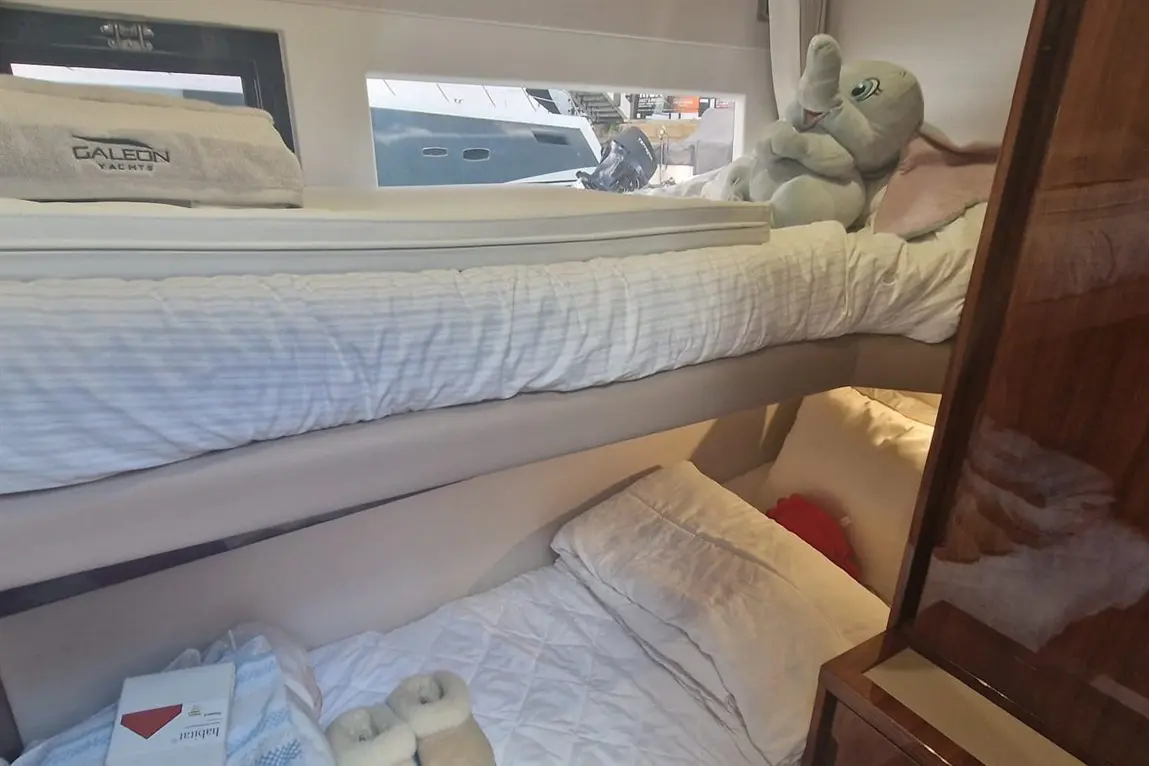 Interior of a yacht cabin with bunk beds, a plush elephant toy, and bedding. A towel with 'GALEON YACHTS' is visible.